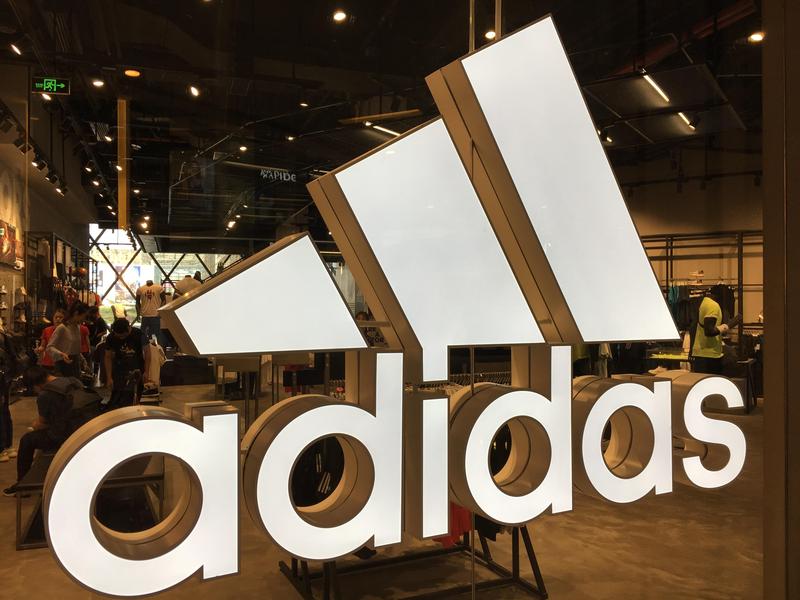 Der Markenschriftzug adidas als leuchtende Profilbuchstaben scheint durch die Schaufensterfront und ist als 3D Werbeschrift prägnant in der Sportswear zu sehen. 