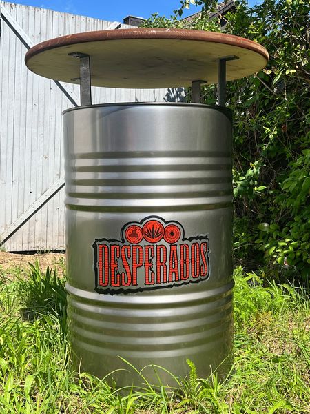 Despsrados-Fasstisch Der Desperados-Fasstisch ist ein robuster Stehtisch aus recyceltem Ölfass mit hochwertiger Douglasien-Holzplatte, ideal für Bars, Beachclubs und Terrassen. Der individuell gebrandete Fass-Stehtisch kombiniert industrielles Design mit sommerlichem Flair und sorgt für ein einzigartiges Gästeerlebnis im Außenbereich.