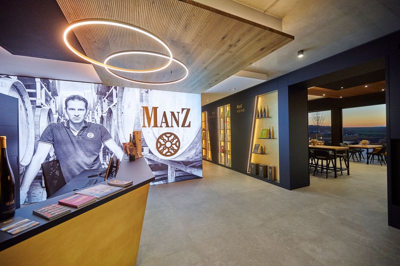 Eine großflächige, hell strahlende Leuchtwand aus Spanntuch zeigt ein Schwarz-Weiß-Motiv aus dem Weinkeller sowie das Manz-Logo am Empfang. In der dunklen Regalwand rechts daneben sind filigrane, hinterleuchtete Schriftzüge aus Acryl direkt in die Elemente durchgesteckt.