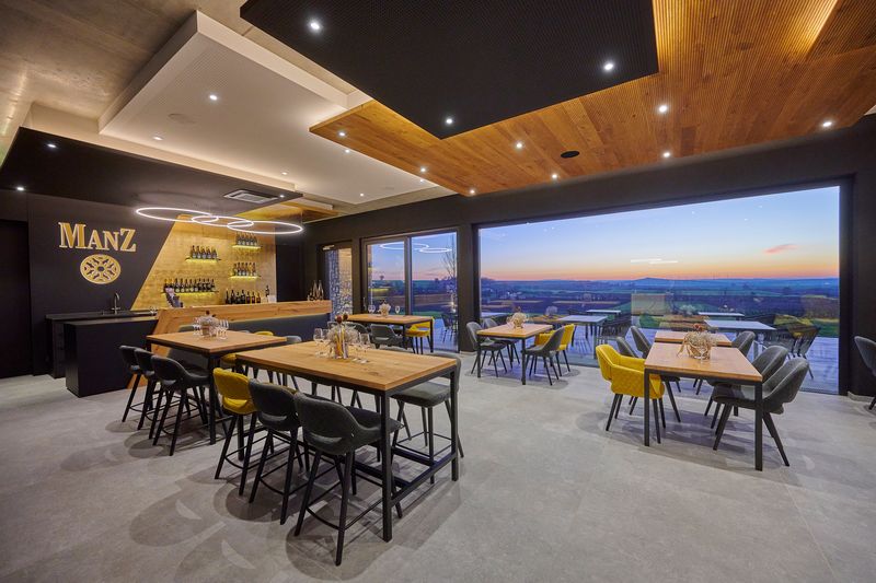 Eine stilvolle Bar, an der das Manz-Logo in Gold effektvoll hinterleuchtet ist. Eine Innenwerbung mit dem  hinterleuchteteten Markenlogo an der Bar und der dezenten Regalbeleuchtung unterstreichen das luxuriöse Ambiente. Perfekt abgestimmte Lichtakzente für eine exklusive Weinpräsentation.