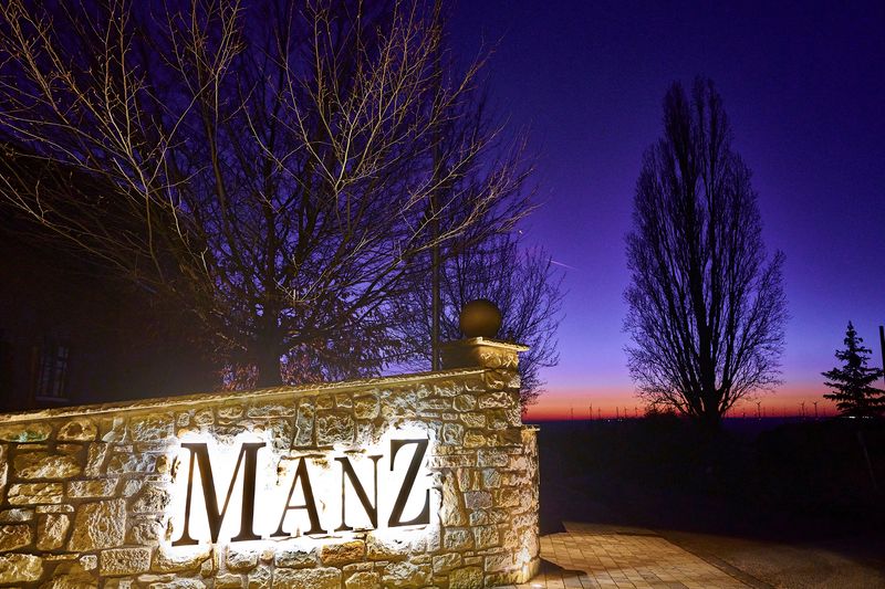 Ein strahlend weiß hinterleuchteter Namenszug „MANZ“ ist direkt auf einer Natursteinmauer am Eingang montiert. Die Beleuchtung am Stein und der starke Kontrast der Lichtbuchstaben ist ein Markanter Wegweiser. Die Lichtwerbung sorgt für Sichbarkeit des Weinguts am rheinhessischen Weinberg.