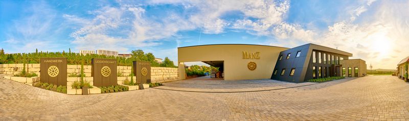 Konzeptuelle Markenpräsenz am Weingut Manz. Die Kombination aus Fassaden-Leuchtwerbung und freistehenden Werbestelen sorgt für ein konsistentes Corporate Design. Das Bild zeigt ein professionelles Branding-Konzept, das Naturstein, dunkle Paneele und edle Metalloptik kombiniert.