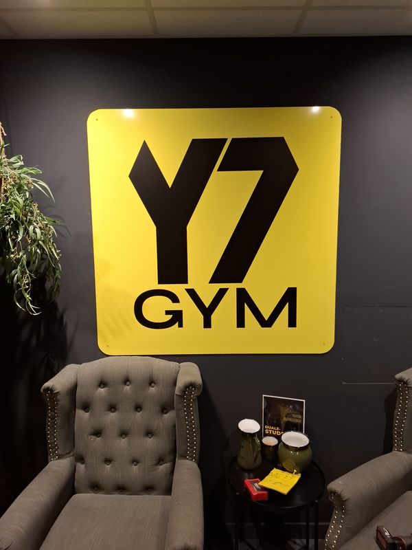 Ein weiteres Highlight ist die Beschilderung in der Lounge in dem Fitnesscenter Y7 Gym. Hier haben wir ein großformatiges, quadratisches Alu-Dibond-Schild installiert. Der starke Kontrast des gelben Schildes zur dunklen Wandgestaltung und den klassischen Polstermöbeln schafft einen modernen, einladenden und professionellen Look.