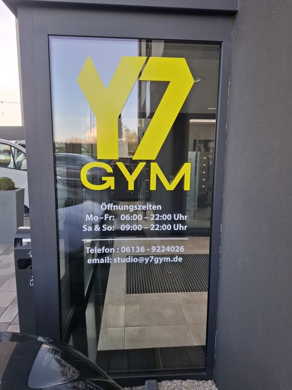 Glasflächenbeschriftung am Eingangsbereich von Y7-GYM. Die Kombination aus großem Y7 GYM Logo in gelber Markenfarbe und präzise geplotteten Informationen wie Öffnungszeiten, Telefonnummer und E-Mail-Adresse sorgt für eine optimale Kundenkommunikation direkt neben dem Eingang.