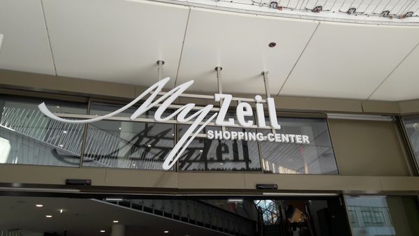 Diese Aufnahme zeigt die Lichtwerbung am Haupteingang des Einkaufscenters. Direkt vor dem Portal-Eingang ist unsere hochwertige Profilschrift des MyZeil-Logos mit der Unterkonstruktioin von der Decke montiert. Diese Beschilderung wirkt frei schwebend und bildet den direkten Empfang für die Kunden.