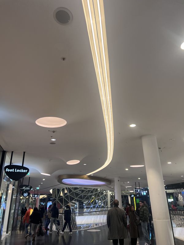 Eindrucksvolle Lichtarchitektur im Innenbereich des MyZeil. Die präzise Montage von geschwungenen LED-Lines, die den organischen Linien der Architektur folgen und für eine homogene Grundbeleuchtung sorgen. Ein besonderes Highlight ist das großformatige, kreisrunde Spanntuch-Lichtelement, das durch energieeffiziente LED-Technik in Violett erstrahlt.