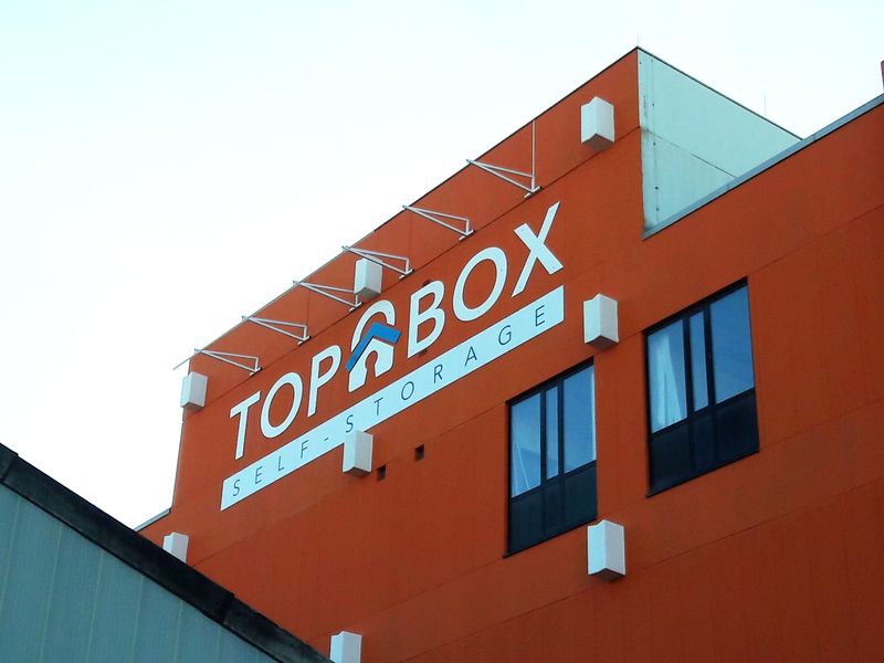 Die Beschriftung ist  eine großformatige Außenwerbung für „TOP BOX SELF-STORAGE“ mit Logo auf einer markanten orangefarbenen Gebäudefassade. In Kombination mit der auffälligen Fassadenfarbe dient die Beschriftung nicht nur als Information, sondern als markantes architektonisches Element. TOP BOX wird durch externe Strahler später von oben perfekt in Szene gesetzt