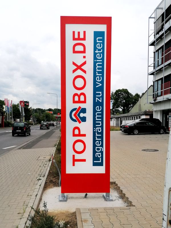 Schlanker, rot-weißer Werbepylon von TO- BOX Self-Storage mit der Aufschrift „Lagerräume zu vermieten“ und Webadresse, montiert auf einem Betonfundament. Der Pylon ist mit energiesparsamen LED-Modulen belegt und leuchtet beidseitig die Acrylfronten aus.