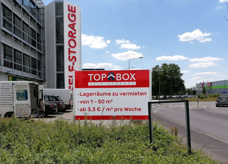 Werbetafel für TOP-BOX Self-Storage zur Vermietung von Lagerräumen, dahinter sieht man den vertikalen roten Leuchtschriftzug am Gebäude.Die informative Preisangaben auf der stabilen Werbetafel mit einer weithin sichtbaren Fassadenbeschriftung vereint. Die freistehende Beschilderung ist beidseitig.