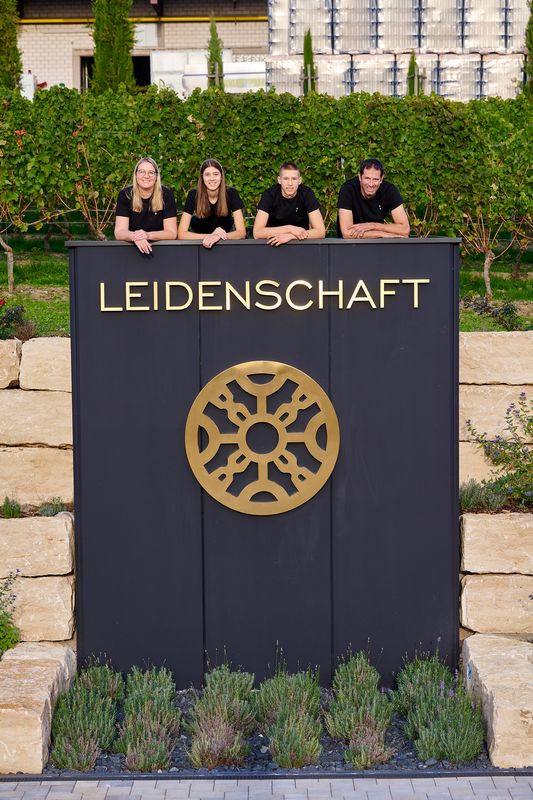 Drei schwarze Stelen leuchten vor einer Natursteinmauer mit den Begriffen „Handwerk“, „Leidenschaft“ und „Faszination“.Jede Einheit enthält ein präzise gefertigtes, golden strahlendes Familienwappen vom Weingut Manz als zentrales Lichtelement.