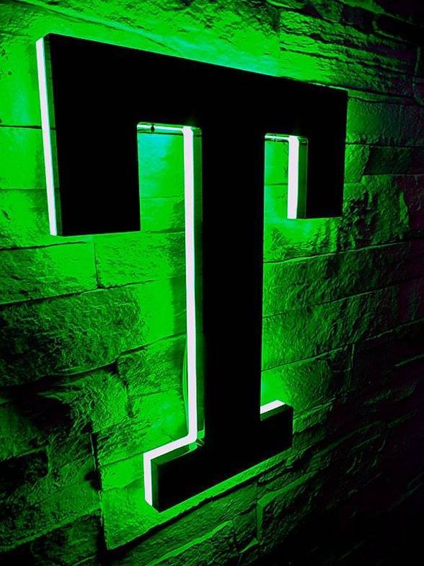 Das T der Letter ist ein Profil 8 Vollacrylleuchtbuchstabe, welches aus gedoppeltem Acrylglas (hinten weiß/Opal vorne schwarz lichtdicht) Front besteht. Die Teilstücke der Zarge bestehen aus lichtdichten Teilen. Die Zargen sind weiß leuchtend und die Rückleuchter machen auf die Wand einen grün leuchtend schimmernden Coronaeffekt.