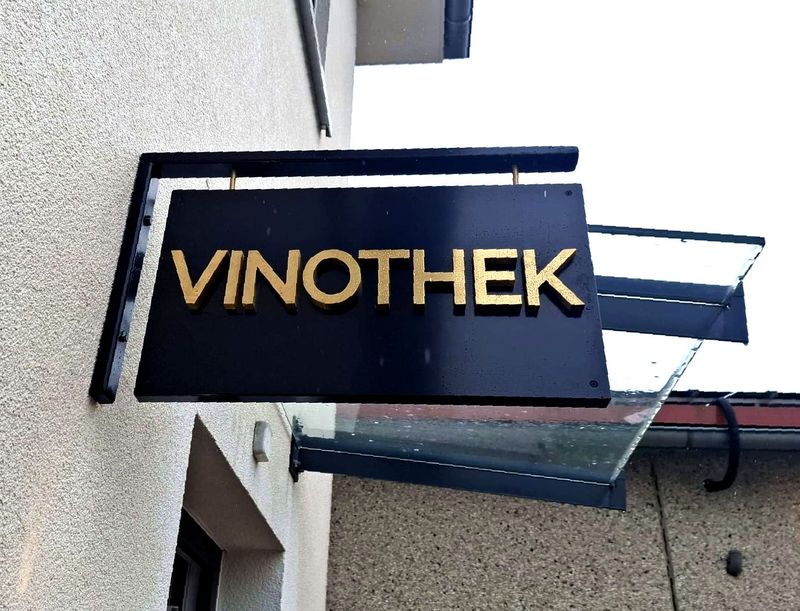 Vergoldeten Reliefbuchstaben mit Rückbeleuchtung Die Beschilderung am Eingang der Vinothek ist als beidseitiges Nasenschild konzipiert und sorgt damit für optimale Sichtbarkeit aus beiden Laufrichtungen. Durch die Kombination aus Metallveredelung und integrierte Rückbeleuchtung heben sich die vergoldeten Buchstaben stilvoll vom Hintergrund ab.