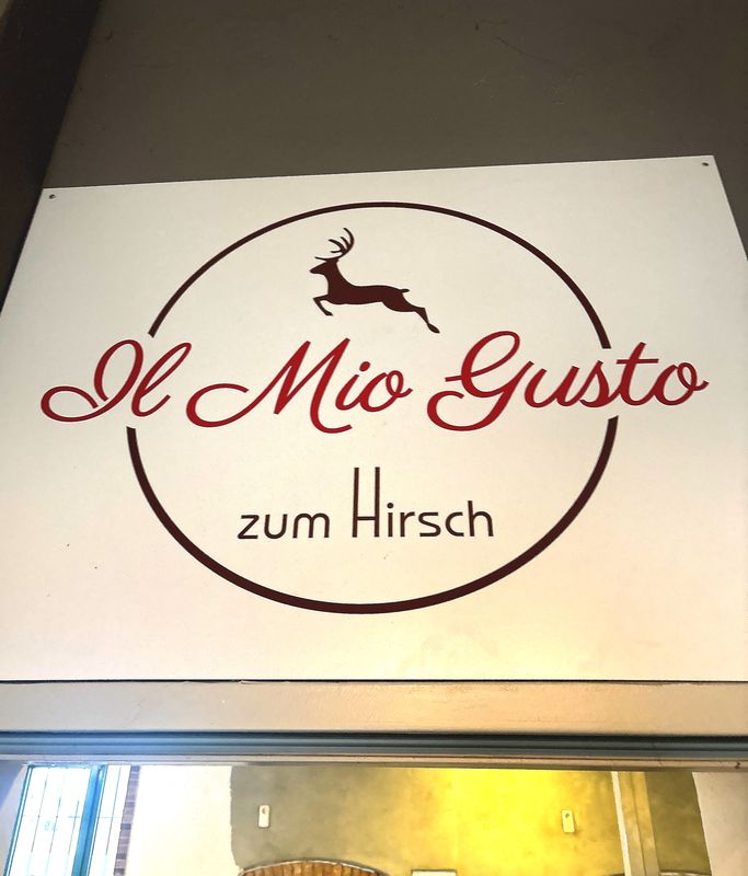 Ein quadratischer leuchtender Indoor-Leuchtkasten mit einer hochwertigen Frontscheibe in Opalweiß. Im Zentrum steht der geschwungene, weinrote Schriftzug „Il Mio Gusto“, der mit Plotterfolie auf die Acrylfront kaschiert wurde. Über einem Türsturz montiert sorgt der Leuchtkasten durch seine homogene LED-Hinterleuchtung für eine einladende Atmosphäre im Eingangsbereich.